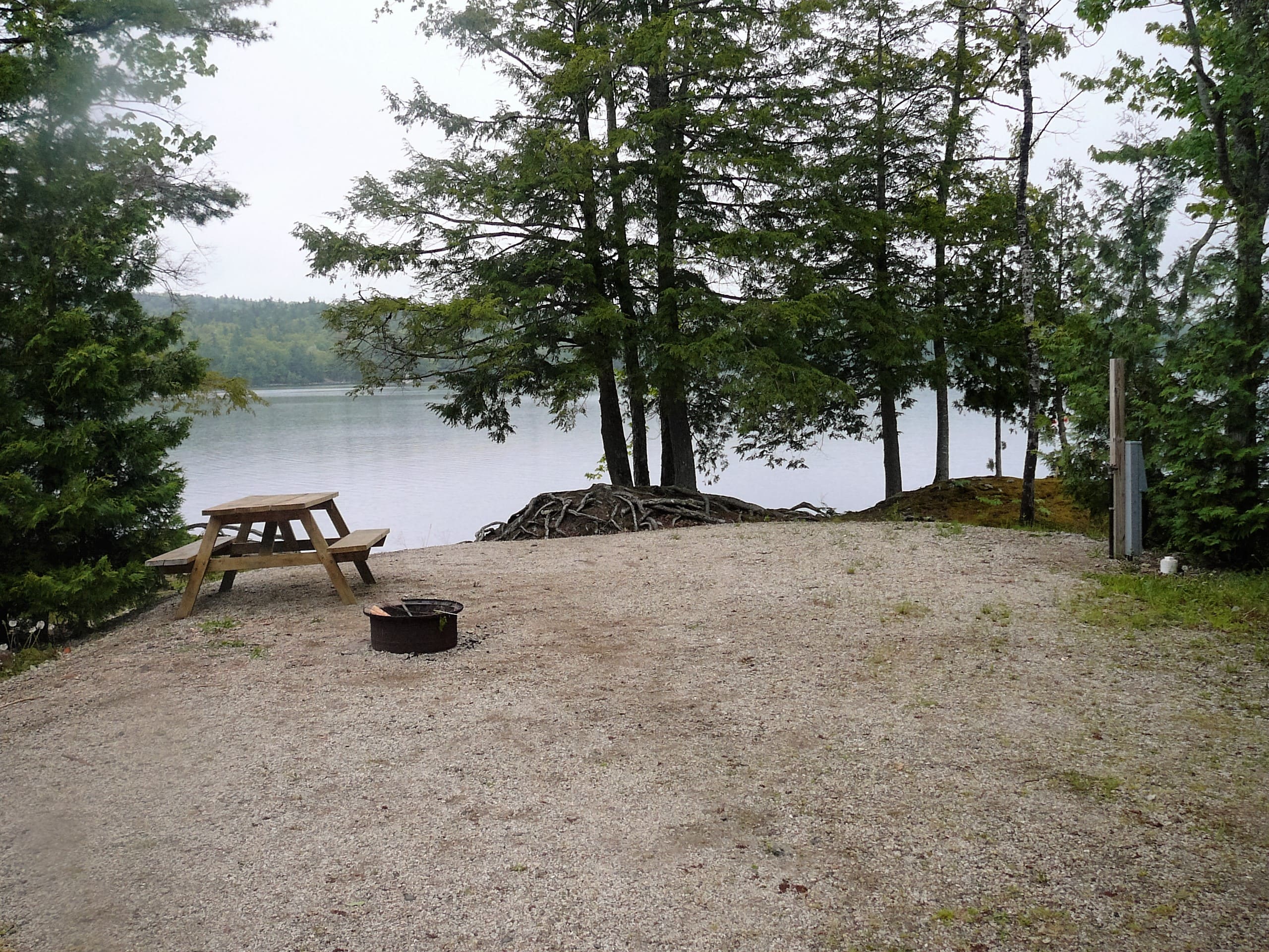 A picnic table and fire pit in the sand near water.